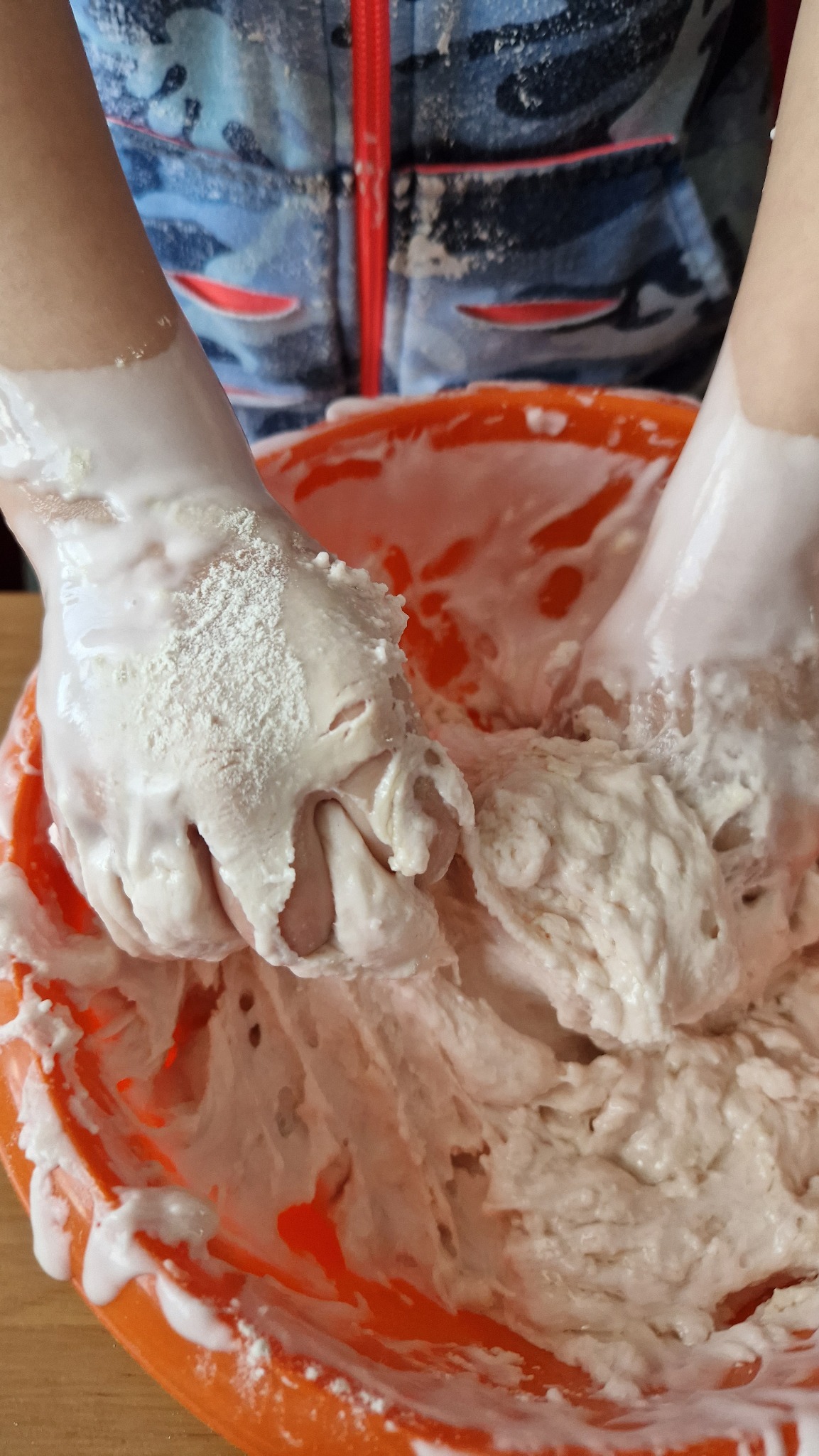 Dwie ręce ugniatają białą masę w pomarańczowej misce, widoczne ślady mąki na dłoniach i ubraniu.