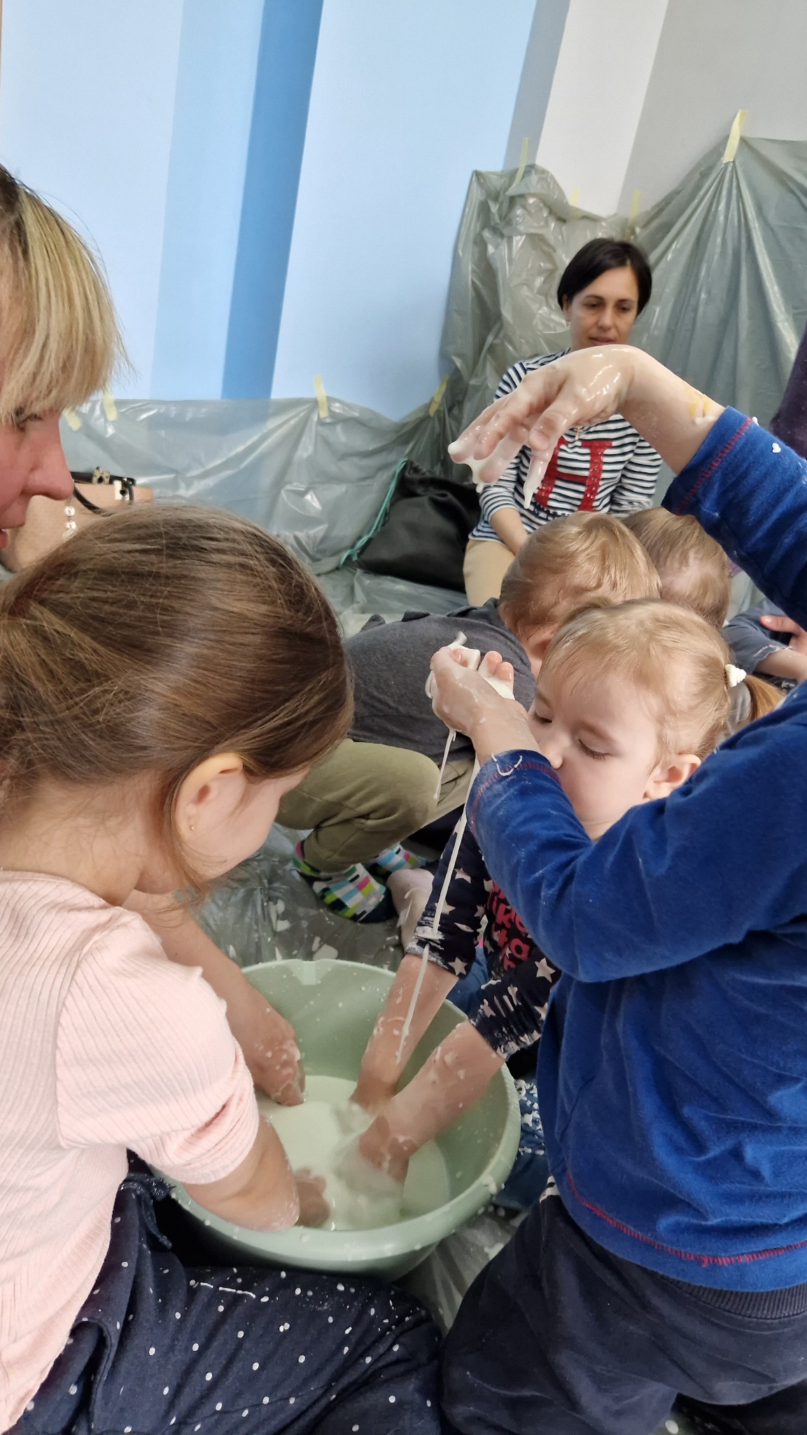 Dzieci bawią się w grupie, zanurzając ręce w misce z białą, lejącą się substancją, podczas zajęć sensorycznych pod nadzorem dorosłych. Tło zabezpieczone folią.