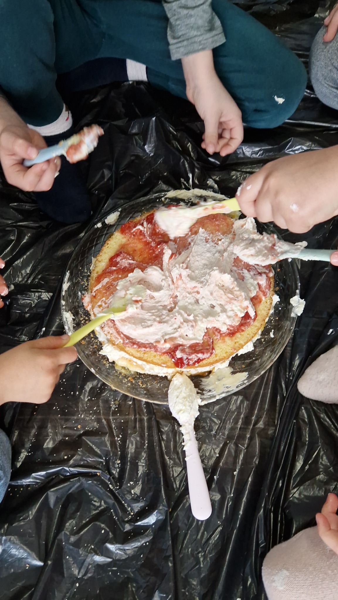 Dzieci dekorują tort na talerzu, używając kolorowych szpatułek, na czarnym plastikowym podłożu.