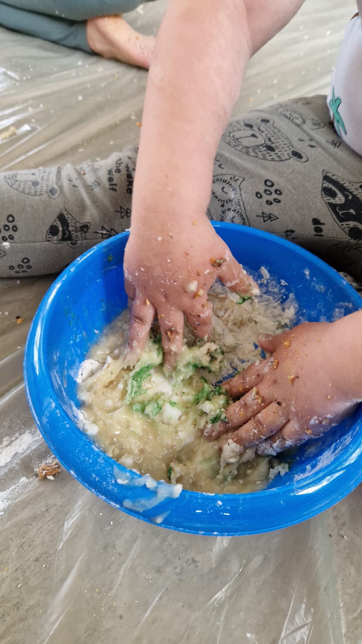 Dziecięce ręce ugniatają masę w niebieskiej misce, prawdopodobnie podczas warsztatów kulinarnych dla dzieci. Masa zawiera zielone elementy.