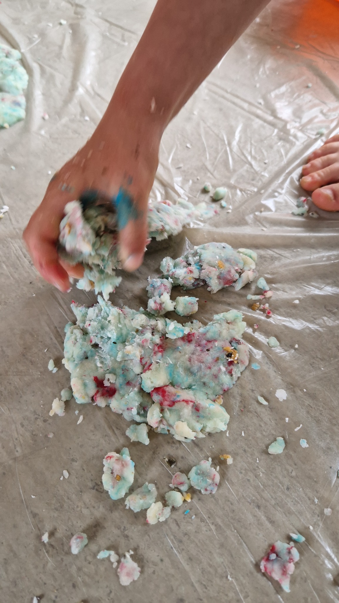 Dziecięca dłoń ugniata kolorową masę plastyczną na folii ochronnej, widoczne ślady niebieskiej farby na palcach i fragment stopy w tle.