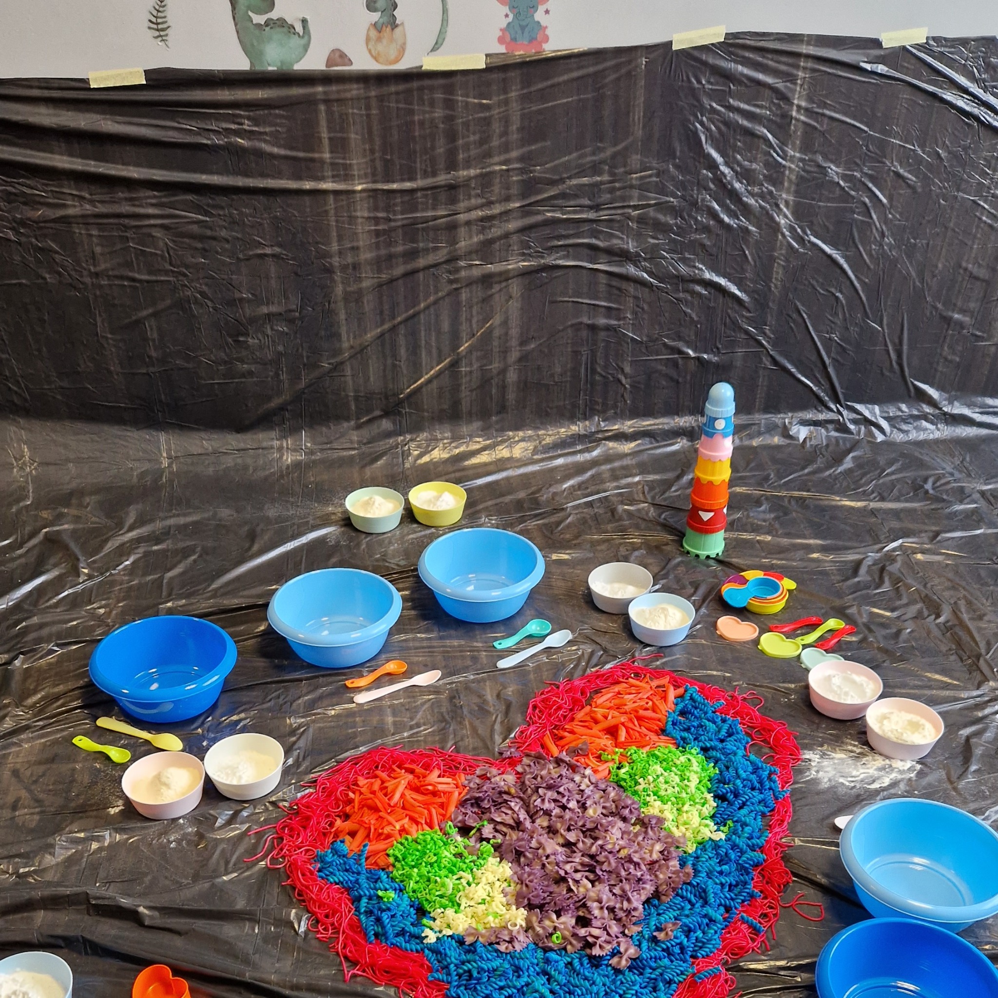 Przygotowane stanowisko do sensorycznej zabawy dla dzieci: kolorowy makaron ułożony w kształt serca, miseczki z białym proszkiem, plastikowe łyżeczki i kubeczki na czarnej folii, tło z motywem...