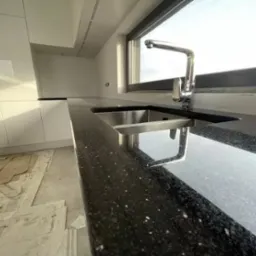 Błyszczący, czarny blat kamienny w kuchni z chromowaną baterią i zlewozmywakiem, widoczne zabezpieczenia podłogi podczas montażu.