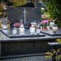 Granity Gdańsk - Polerowany, ciemny granitowy pomnik z białym krzyżem, zniczami i kwiatami na cmentarzu, częściowo zasłonięty przez gałązki z liśćmi.