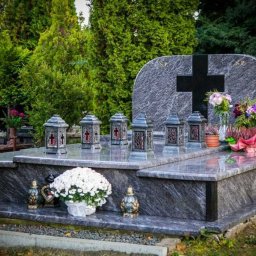 Granity Gdańsk - Granitowy pomnik z czarnym krzyżem, ozdobiony lampionami i kwiatami na cmentarzu, otoczony zielenią drzew.