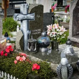 Granitowy nagrobek z krzyżem stylizowanym na drewno, otoczony kwiatami i lampionami na cmentarzu.