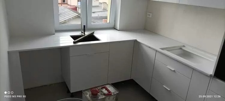 Nowoczesna, biała kuchnia z narożnym blatem, czarnym zlewozmywakiem i widocznym oknem. Na podłodze leży puszka z akcesoriami.