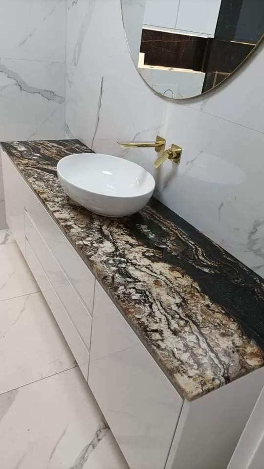 Łazienka z blatem o unikalnym wzorze kamienia w odcieniach brązu, czerni i bieli, umywalką nablatową i złotą armaturą na tle marmurowych płytek.