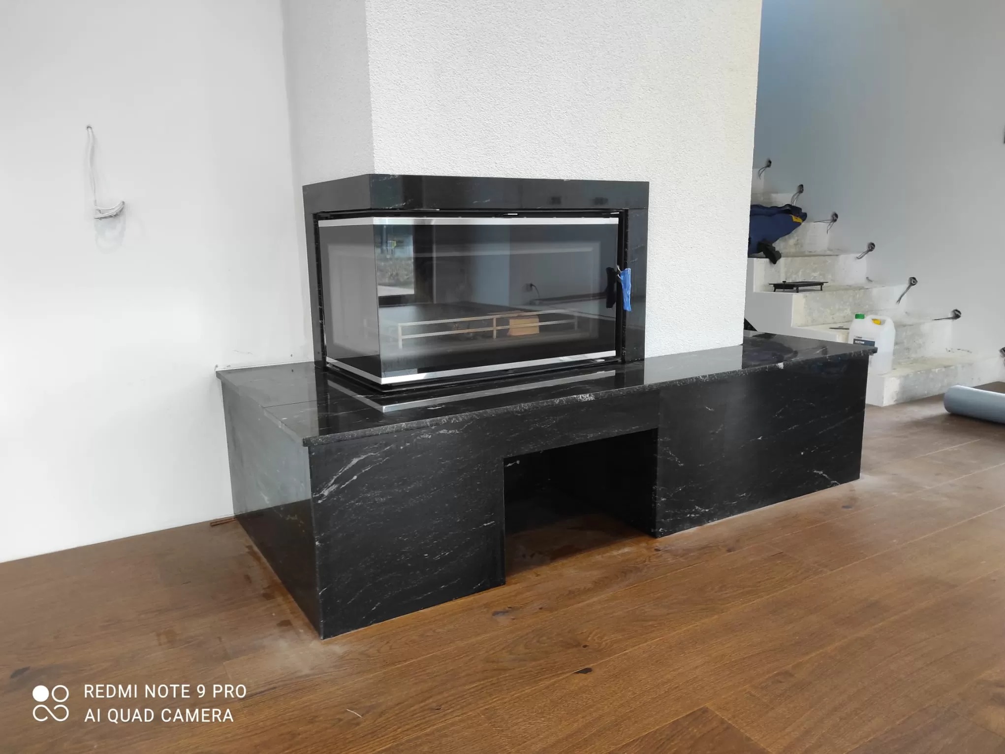 Czarny, marmurkowy obudowany kominek z przeszklonym paleniskiem, na drewnianej podłodze, w surowym wnętrzu z niedokończonymi schodami.