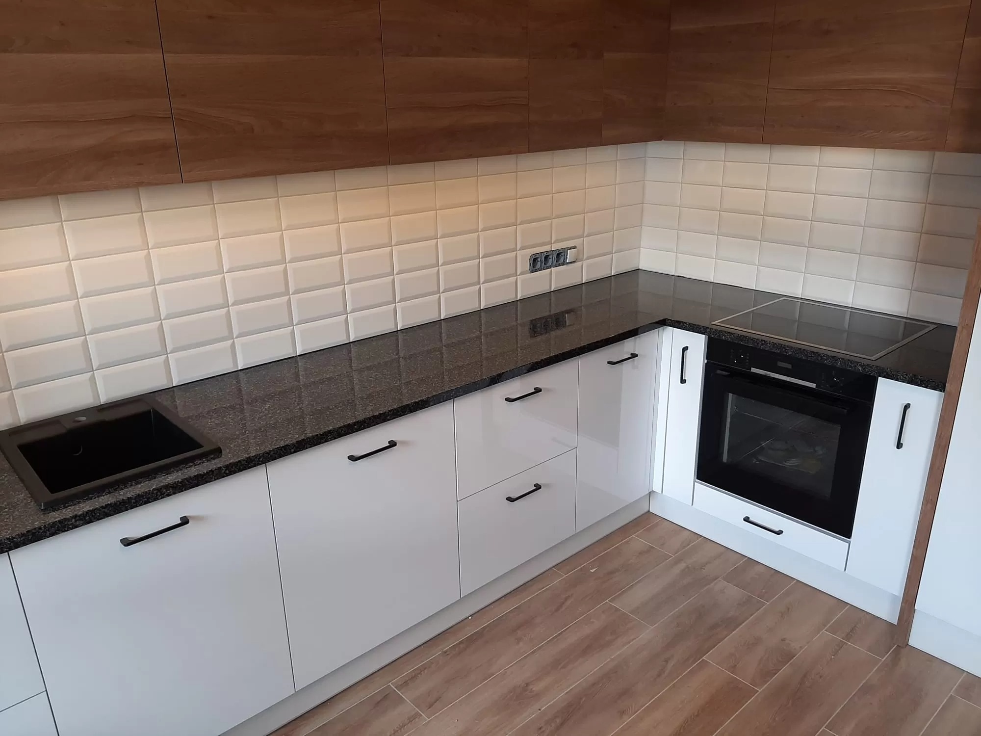 Kąt ujęcia na nowoczesną kuchnię z ciemnym blatem z widoczną fakturą, białymi szafkami z czarnymi uchwytami, białymi płytkami i drewnianymi górnymi szafkami.