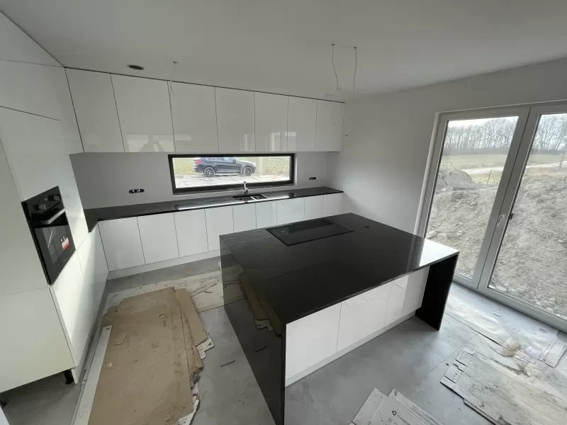 Nowoczesna kuchnia z białymi szafkami i czarnym blatem wyspy kuchennej, widok na zewnątrz przez okno, surowy stan wykończenia podłogi.