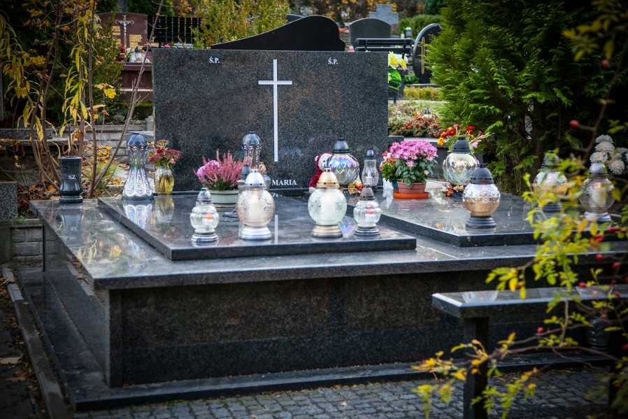 Polerowany, ciemny granitowy pomnik z białym krzyżem, zniczami i kwiatami na cmentarzu, częściowo zasłonięty przez gałązki z liśćmi.