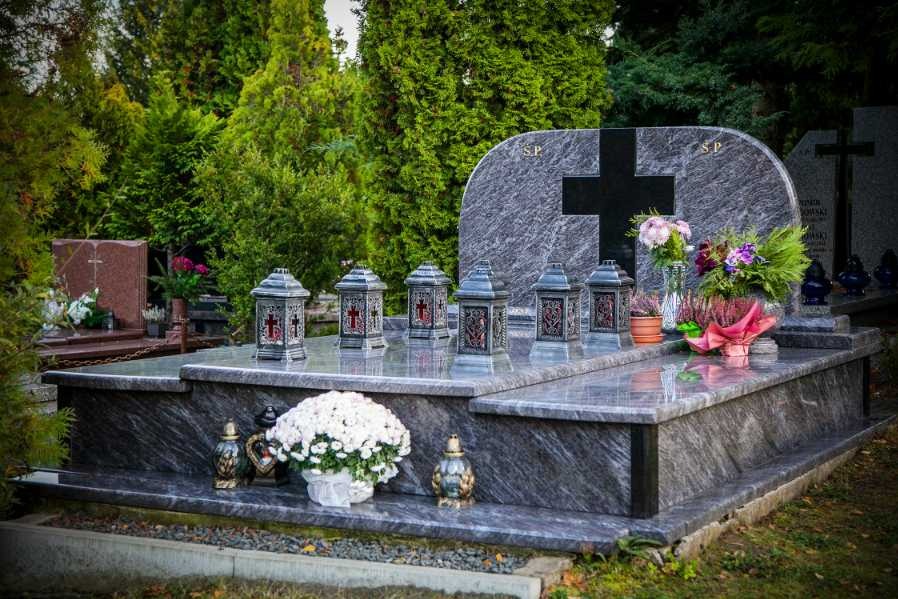 Granitowy pomnik z czarnym krzyżem, ozdobiony lampionami i kwiatami na cmentarzu, otoczony zielenią drzew.