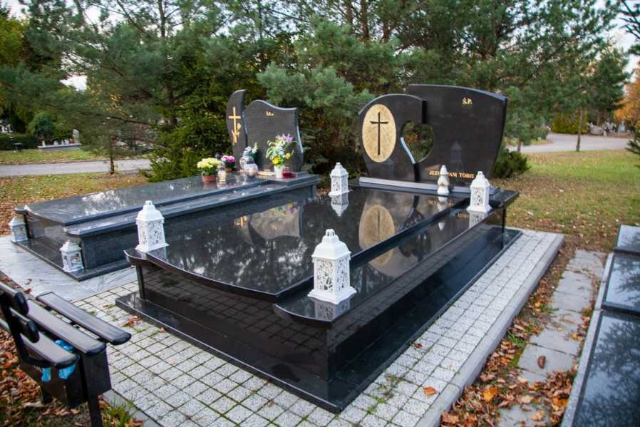 Dwa czarne, polerowane pomniki na cmentarzu, ozdobione kwiatami i lampionami, z refleksami nieba i drzew na powierzchni kamienia.