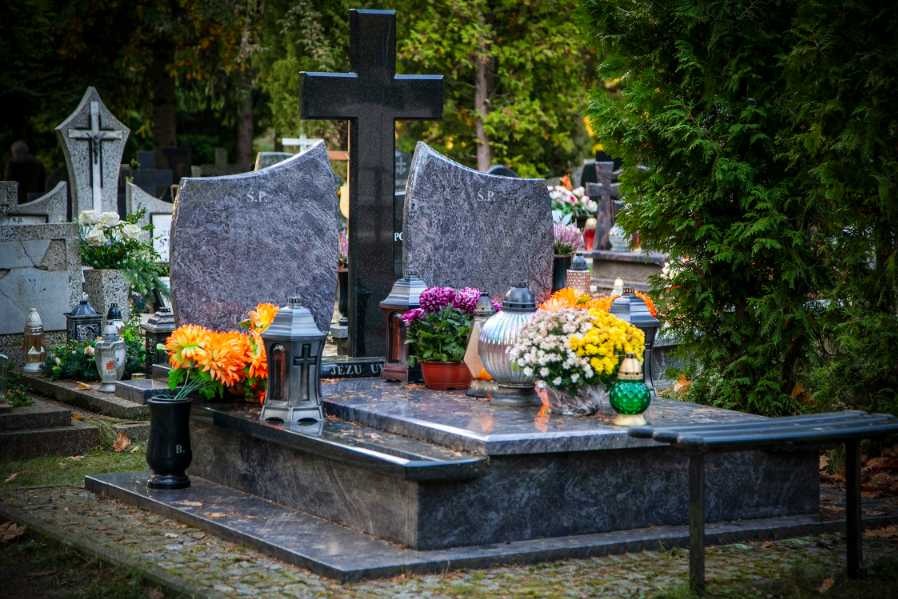 Granitowy pomnik z ciemnym krzyżem, ozdobiony świeżymi kwiatami i lampionami na cmentarzu, w tle zielona roślinność.