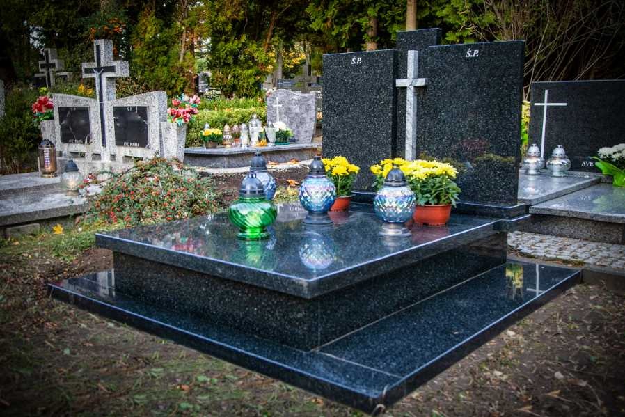 Czarny, polerowany pomnik z trzema lampionami i doniczkami z żółtymi kwiatami na płycie nagrobnej, na cmentarzu z innymi nagrobkami w tle.