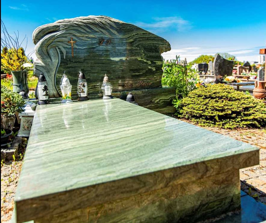 Zielony nagrobek z falistym wzorem kamienia i krzyżem, ozdobiony zniczami, na tle cmentarza z krzewami i innymi grobami.