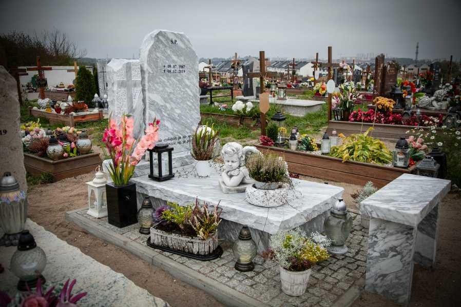 Jasny, marmurowy pomnik z inskrypcją i datami, ozdobiony figurką anioła, kwiatami w doniczkach, lampionami i zniczami, na cmentarzu z widokiem na zabudowania mieszkalne.