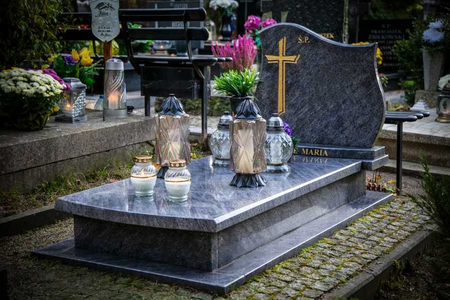 Granitowy pomnik z inskrypcją i złotym krzyżem, ozdobiony szklanymi lampionami i świecami, otoczony zielenią cmentarza.