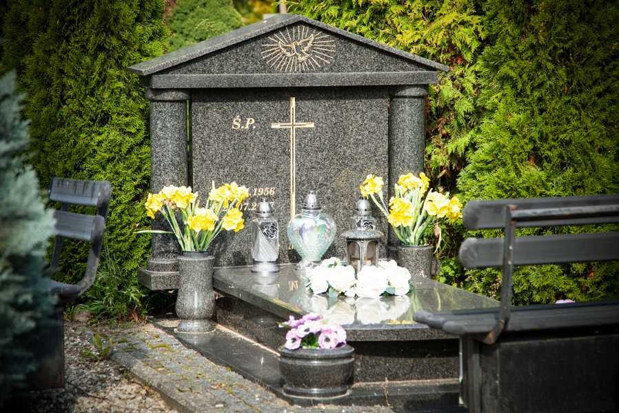 Granitowy pomnik z wyrytym krzyżem i gołębiem, ozdobiony świeżymi żółtymi narcyzami w wazonach i lampionami, otoczony zielonymi tujami i ławkami.