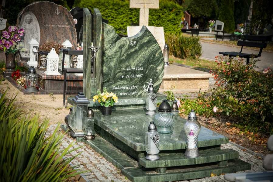Zielony, marmurkowy pomnik z metalowym krucyfiksem i wygrawerowanymi datami, otoczony lampionami i kwiatami na cmentarzu.