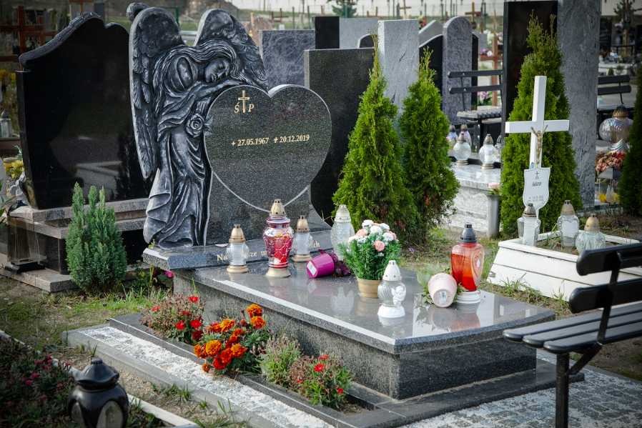 Granitowy pomnik z rzeźbą anioła, sercem z datami i krzyżem, otoczony lampionami, kwiatami i iglakami na cmentarzu.