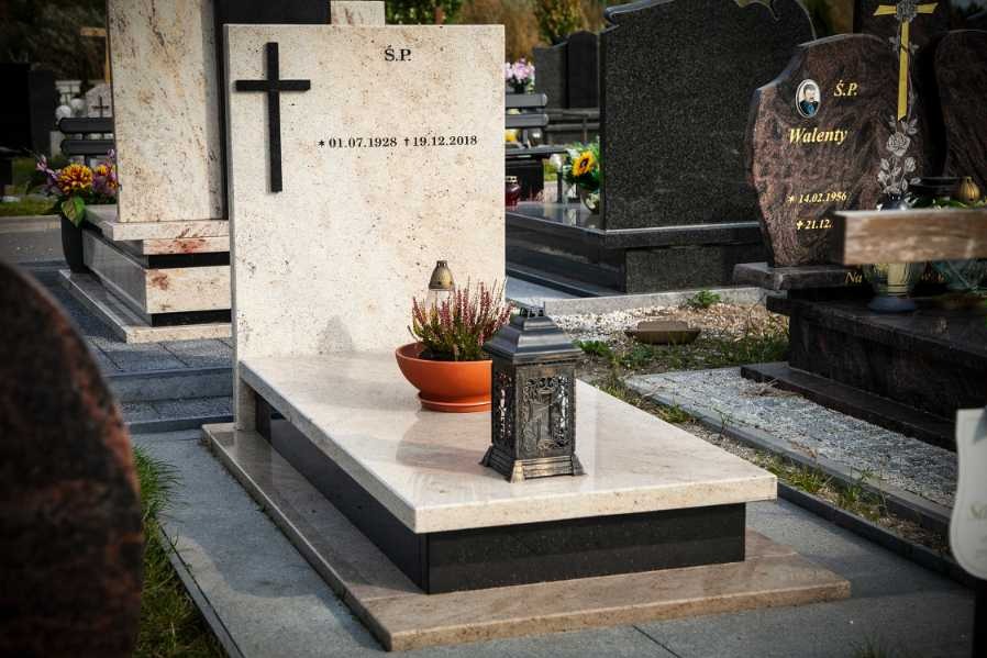Jasny, beżowy nagrobek z czarnym krzyżem, datami i napisem 'S.P.', zdobiony rośliną w doniczce i metalowym lampionem, na tle innych nagrobków na cmentarzu.