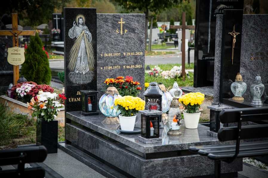 Granitowy pomnik z wizerunkiem Jezusa Miłosiernego i kwiatami, otoczony lampionami i zniczami na cmentarzu.