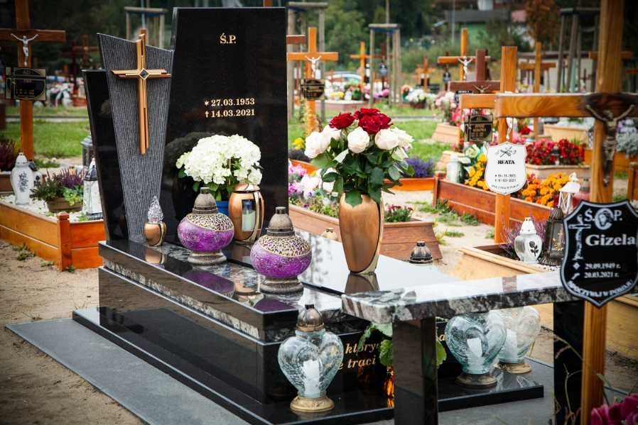 Czarny, polerowany pomnik z krzyżem i datami na cmentarzu w Gdańsku, ozdobiony lampionami i kwiatami w odcieniach bieli i czerwieni.