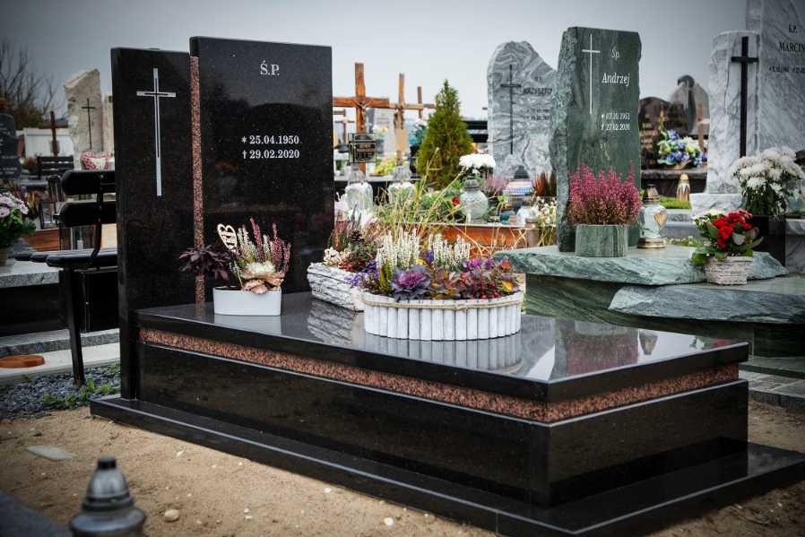 Czarny, polerowany pomnik z granitu z wygrawerowanym krzyżem i datami, udekorowany kompozycjami kwiatowymi na cmentarzu, w tle inne nagrobki.
