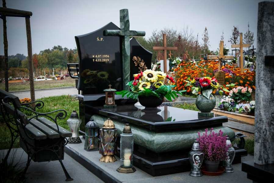 Czarny nagrobek z zielonym krzyżem i datami, ozdobiony kwiatami, z ławką obok, na tle cmentarza z innymi nagrobkami i krzyżami.