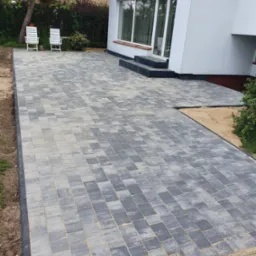 Ułożona kostka brukowa w odcieniach szarości na podwórku przed białym domem z widocznymi schodami i dwoma białymi krzesłami ogrodowymi w tle.