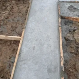 Świeżo wylany beton w szalunku z desek, widoczna ziemia wokół, etap budowy.