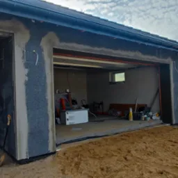 Surowy stan domu w trakcie prac: ściany z szarego styropianu, okno zabezpieczone folią, widoczny wjazd do garażu, piasek na posesji, w tle drugi dom i ż&oacute;łty przew&oacute;d gazowy.