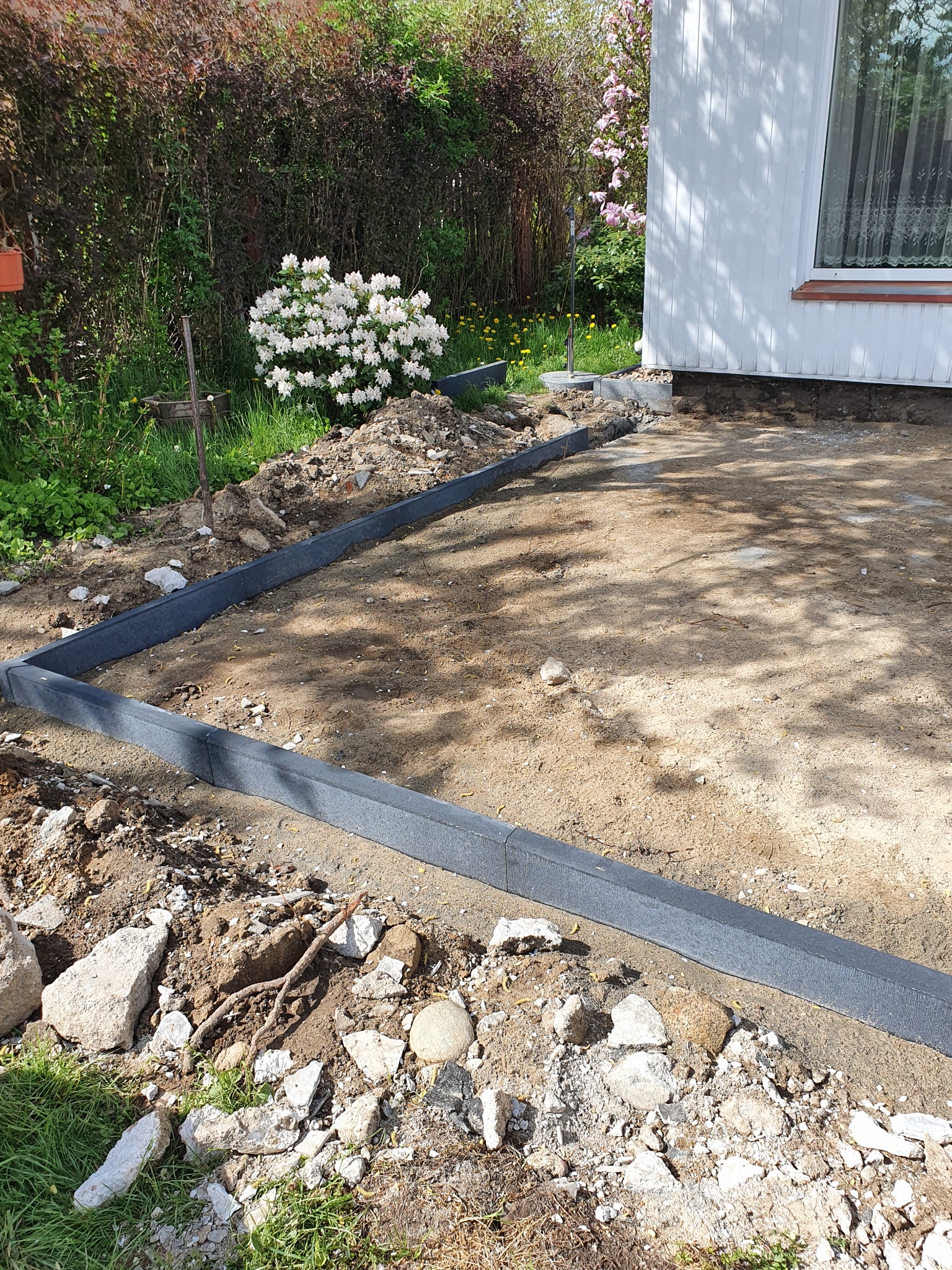 Przygotowanie terenu pod wylewkę fundamentów: Ułożone szalunki z szarego tworzywa, widoczne wykopane podłoże, gruz i roślinność.