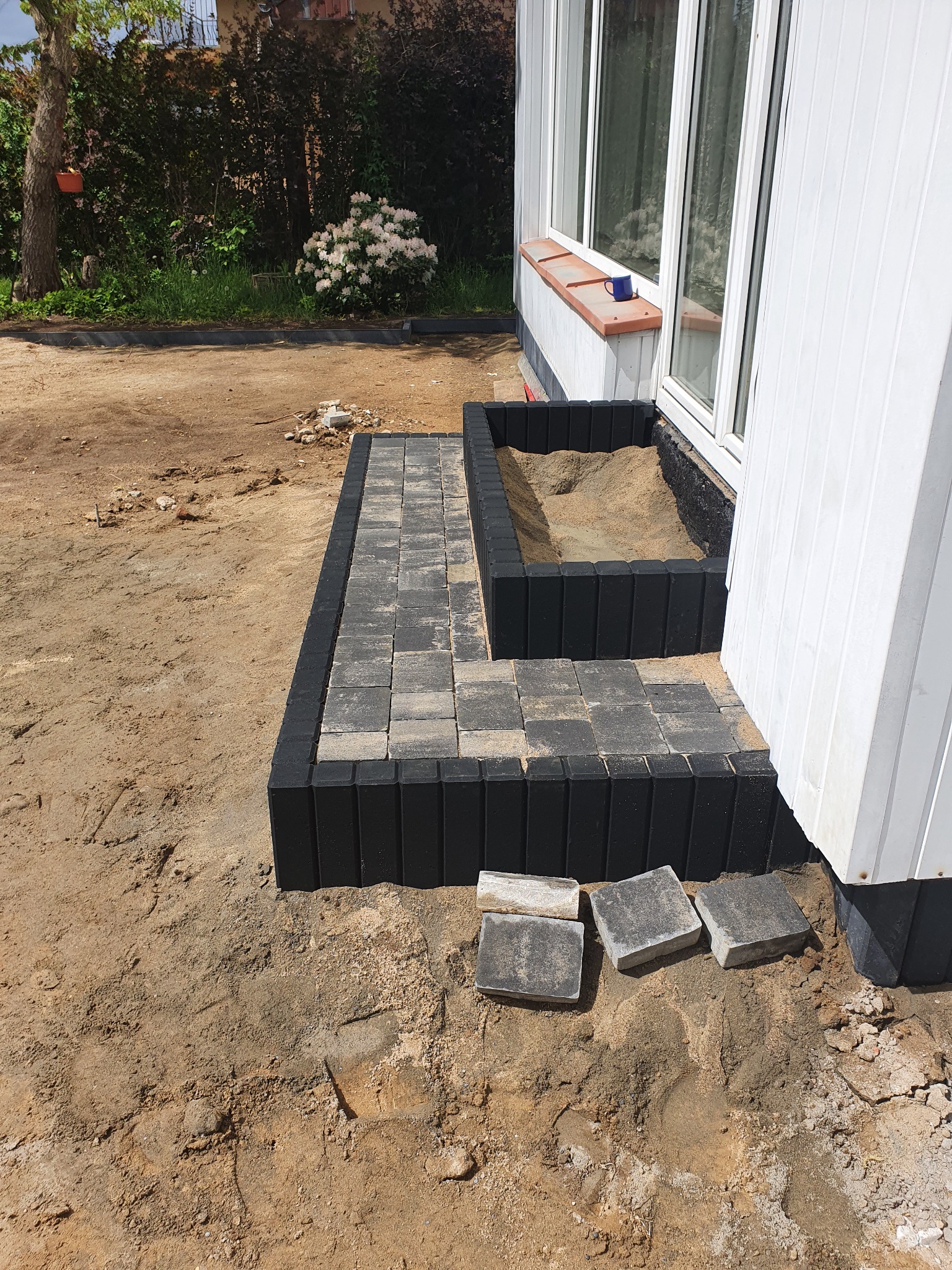 Układanie kostki brukowej przy białej elewacji domu, widoczny czarny obrzeże i piasek, trzy kostki brukowe leżące obok.