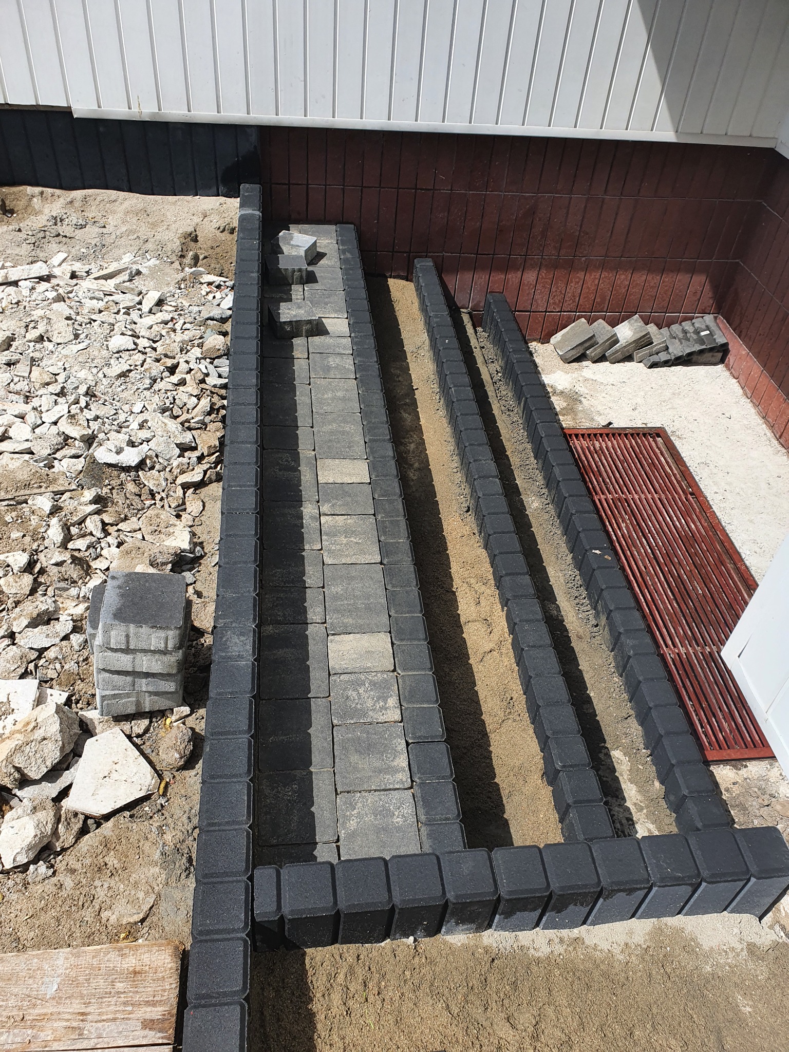 Wykonywanie obrzeży z kostki brukowej wokół budynku, widoczny etap prac z piaskiem wewnątrz koryta, gruz i płyty obok.