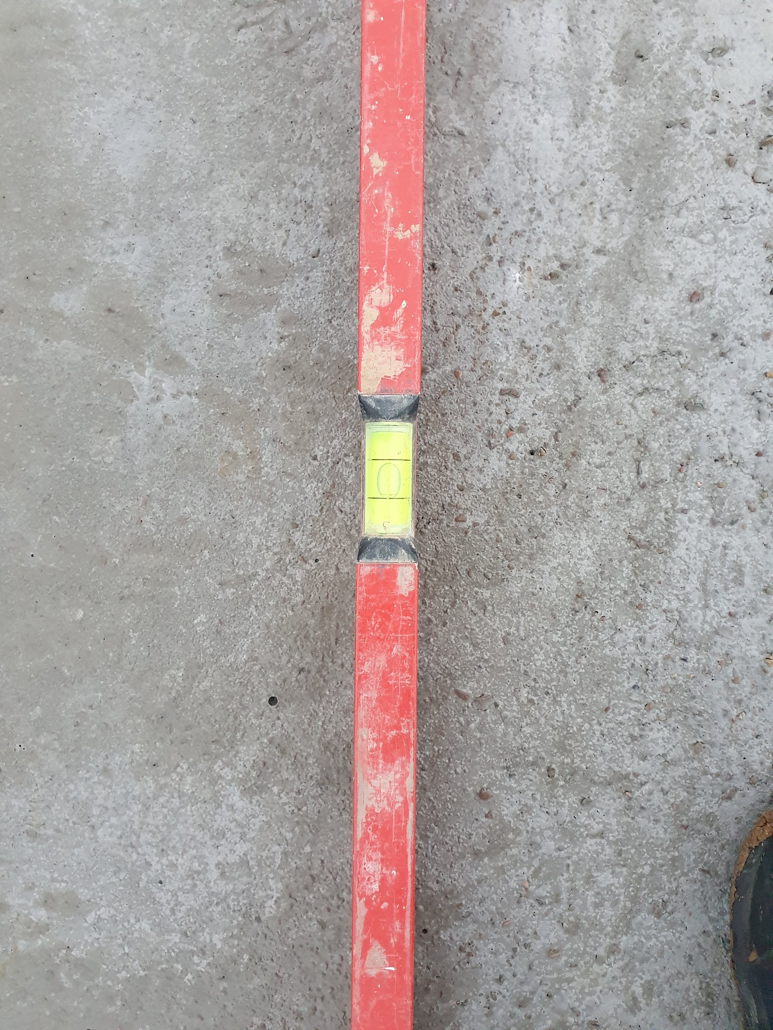 Poziomica w kolorze czerwonym, ułożona na szarej, chropowatej powierzchni betonowej, z widocznym pęcherzykiem powietrza wskazującym poziom.