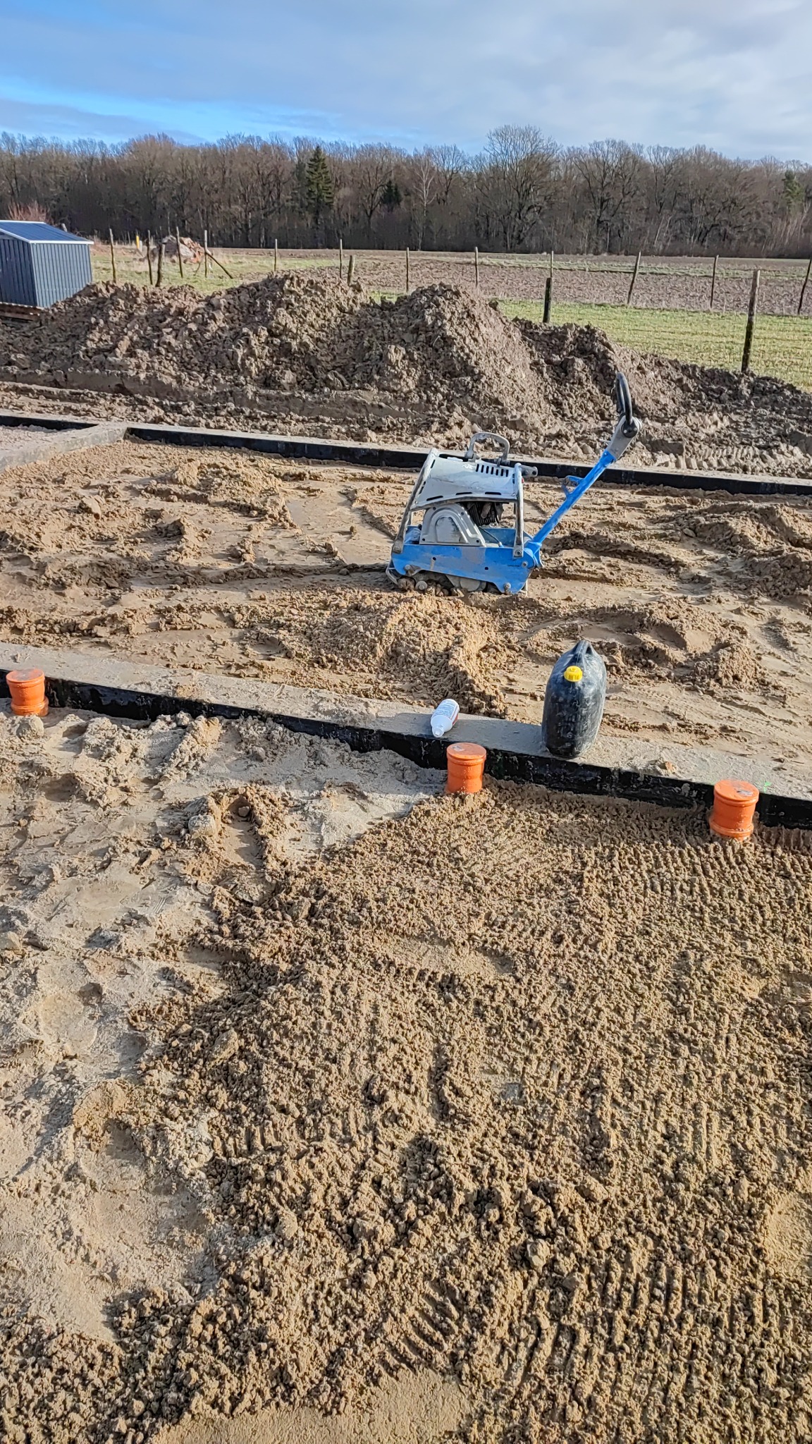 Przygotowanie terenu pod fundamenty: ubijanie piasku zagęszczarką w wykopie z czarną folią, widoczne pomarańczowe znaczniki, pryzma ziemi w tle.