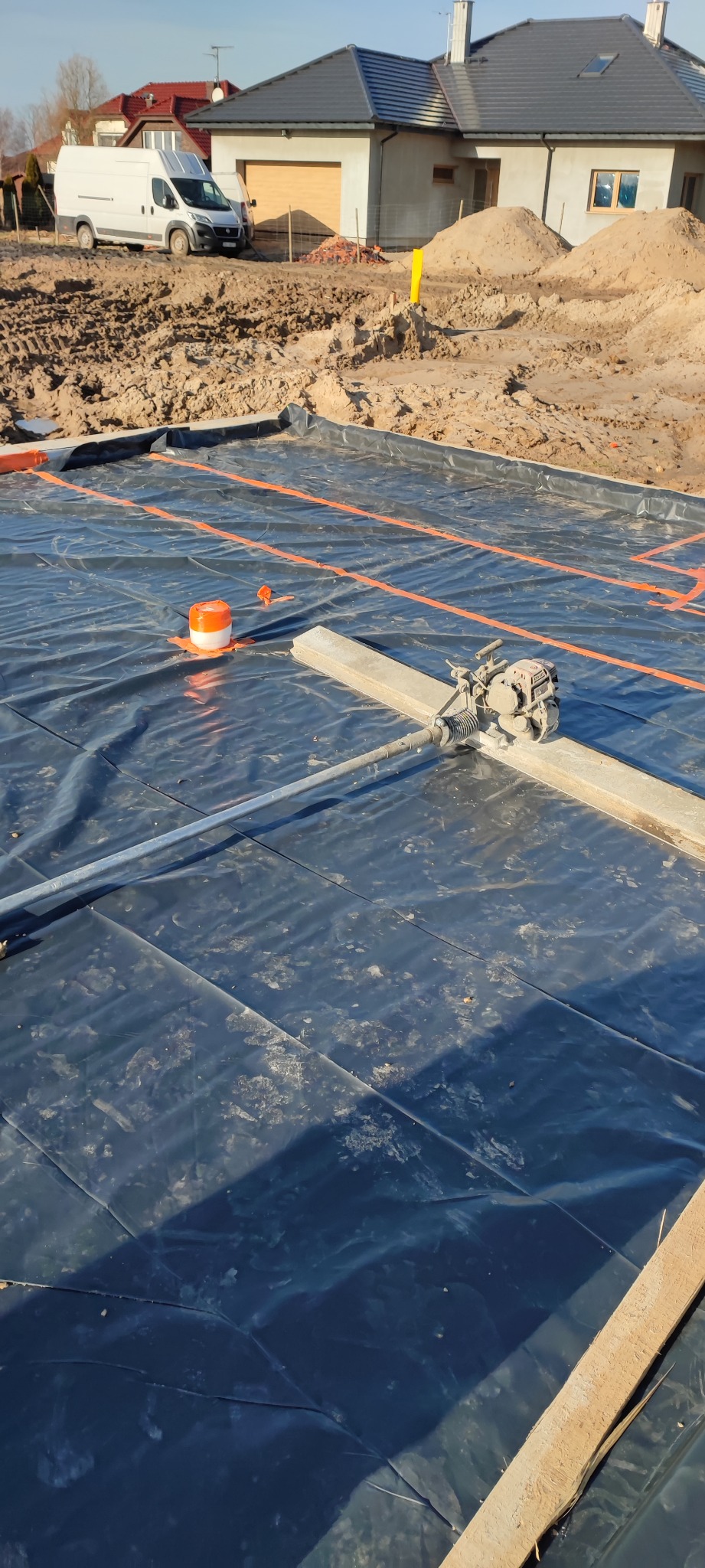 Przygotowanie podłoża pod fundamenty w Białogardzie: czarna folia hydroizolacyjna z pomarańczowymi oznaczeniami i ubijarką wibracyjną na drewnianej łacie, w tle budowa domu jednorodzinnego.