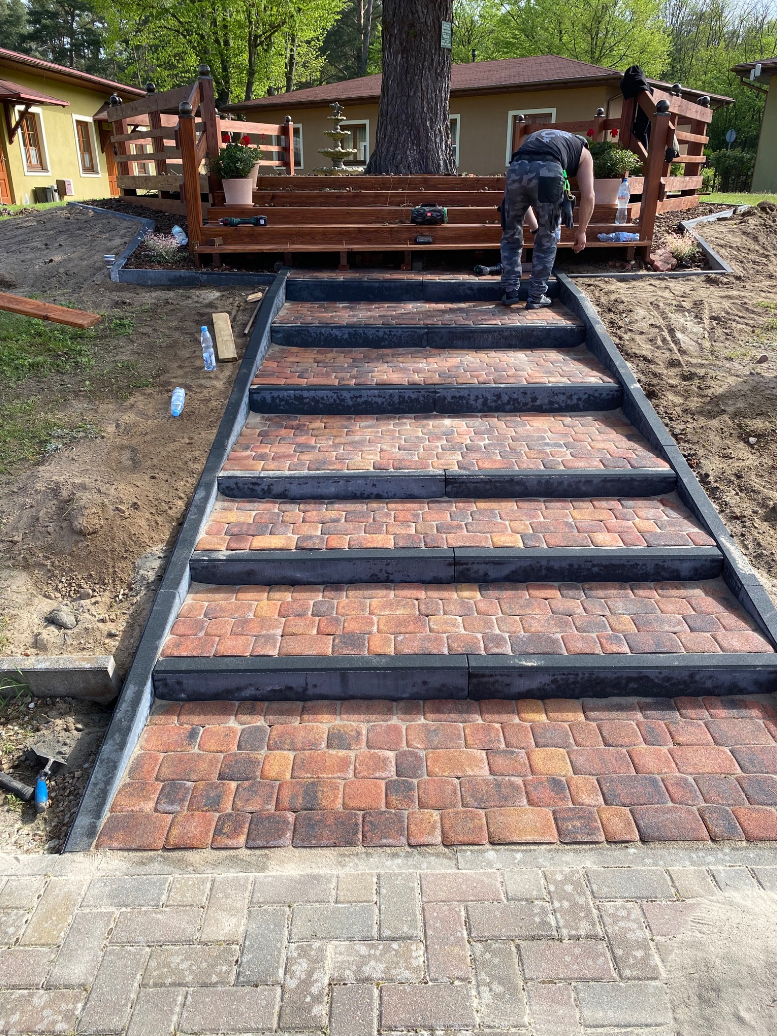 Schody z kostki brukowej prowadzące do drewnianego tarasu, wykonawca w trakcie pracy, widoczne narzędzia i butelki z wodą.