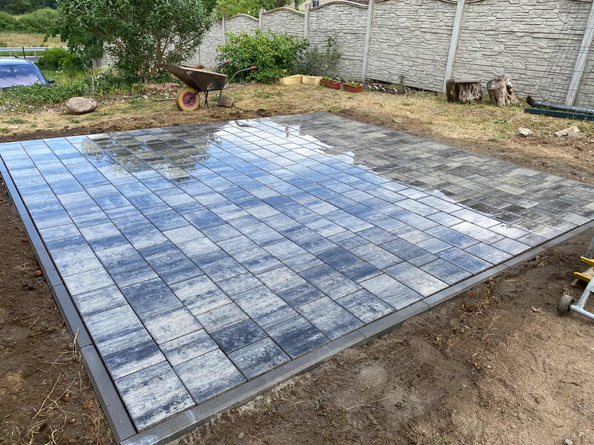 Częściowo ułożona, szara kostka brukowa na posesji, z widoczną taczką i fragmentem ogrodzenia w tle, w trakcie prac wykończeniowych.