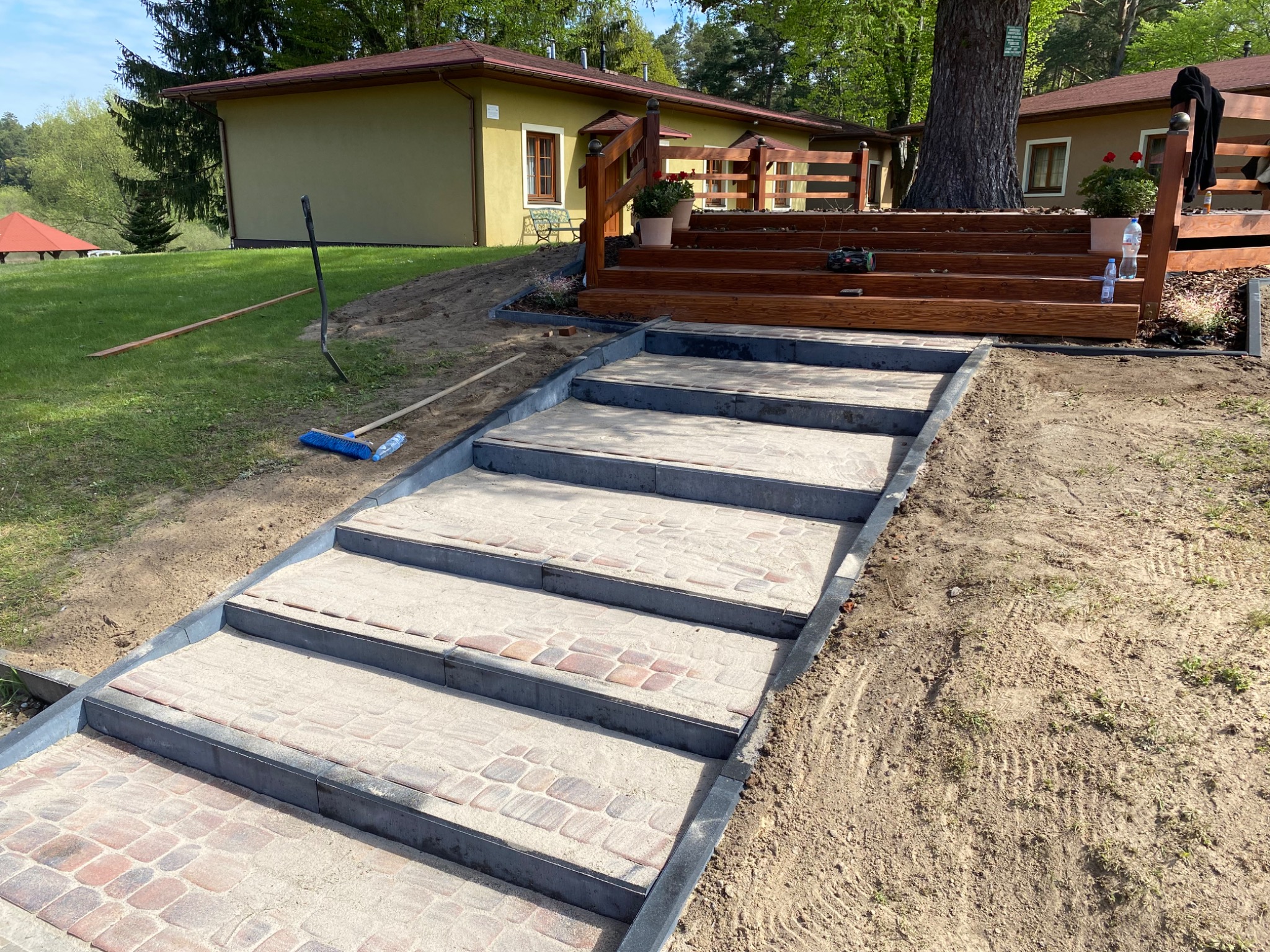 Schody z kostki brukowej prowadzące do drewnianego tarasu z balustradą, widoczne budynki w tle, teren w trakcie prac wykończeniowych.