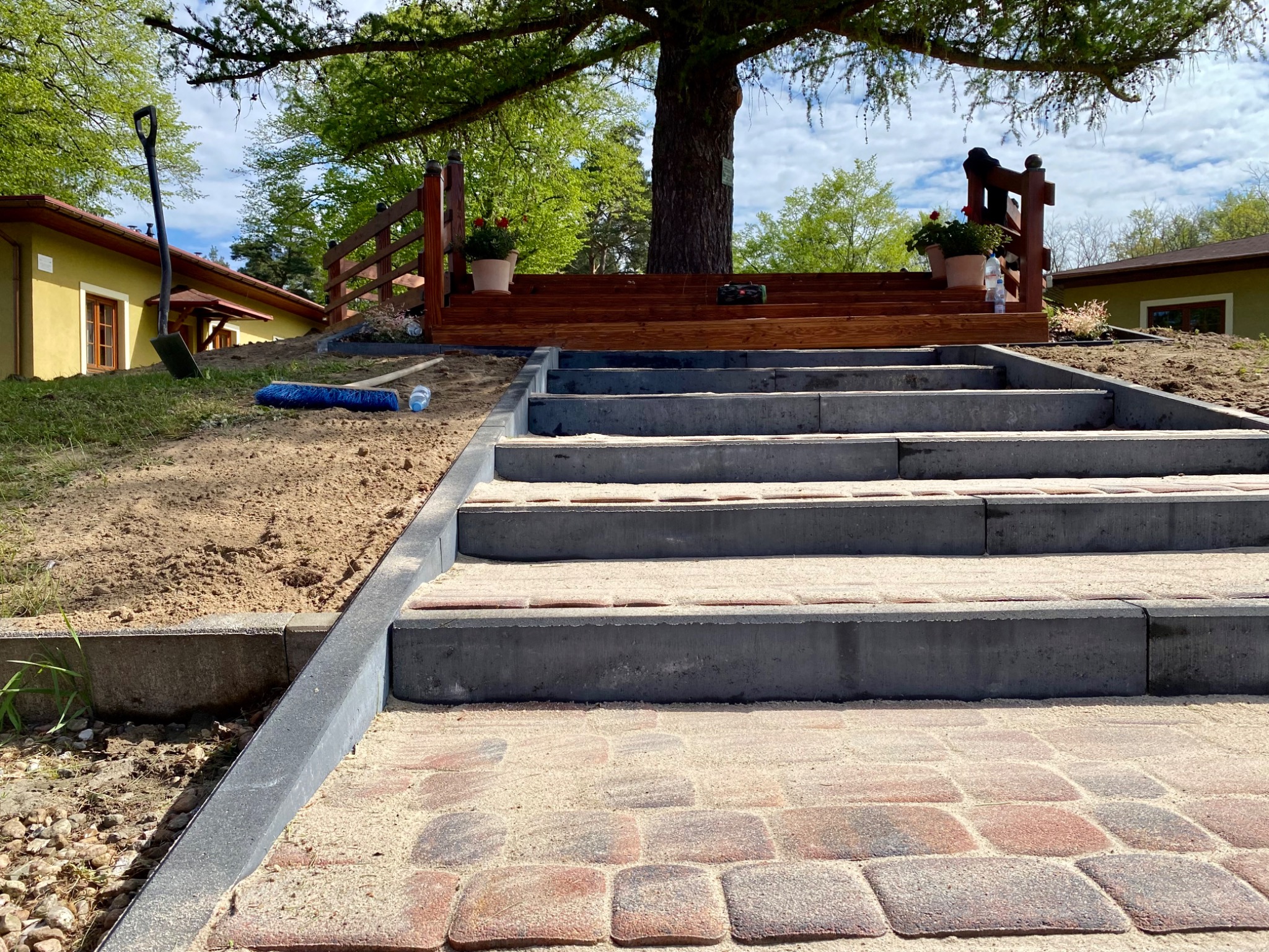 Nowo ułożona kostka brukowa prowadząca do drewnianego podestu z balustradą i kwiatami w donicach, widok z dołu, z boku widoczna łopata i miotła.