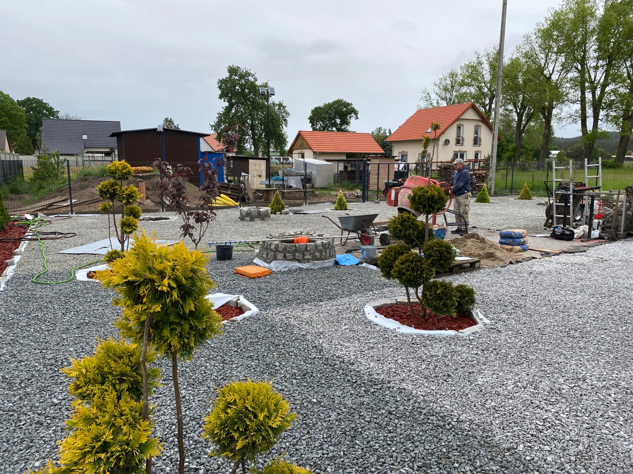 Układanie szarej kostki brukowej na posesji z widocznym pracownikiem przy betoniarce, przygotowanymi workami cementu, paletą z kostką, w tle domy jednorodzinne i elementy dekoracyjne w postaci...