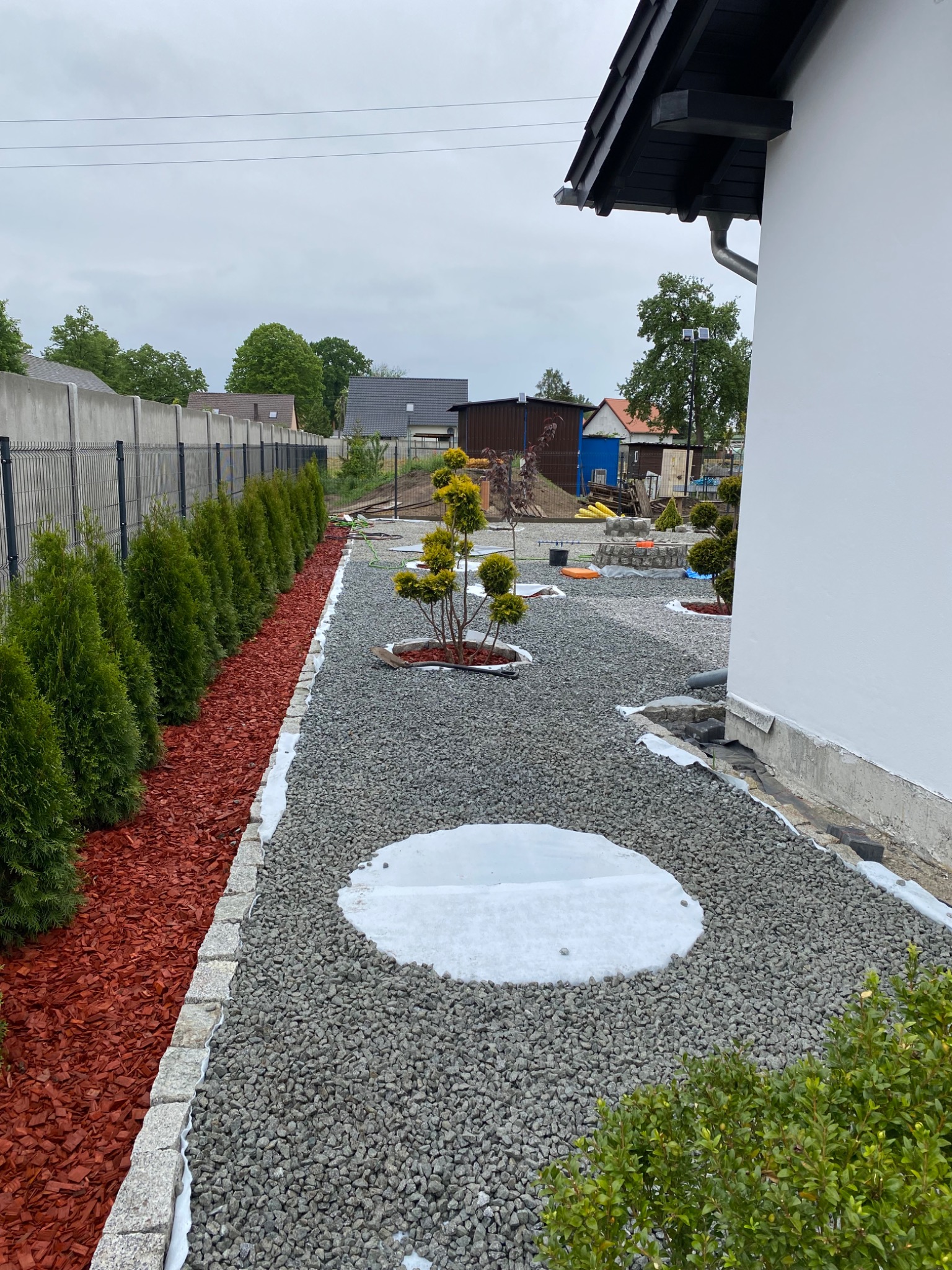 Ścieżka z szarego żwiru wzdłuż białego domu, obrzeża z kostki granitowej, tuje i czerwona kora po lewej stronie, widok na ogród w pochmurny dzień.