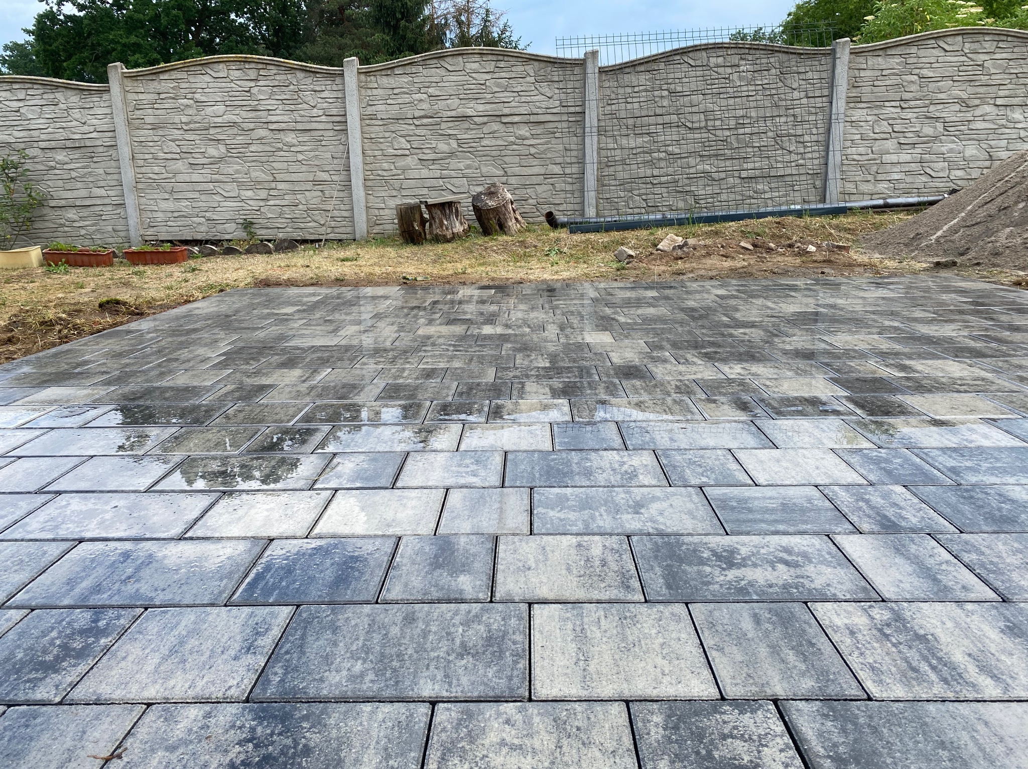 Świeżo ułożona, mokra kostka brukowa w odcieniach szarości, z widocznymi refleksami nieba, na tle betonowego ogrodzenia i przygotowanego podłoża z piaskiem.