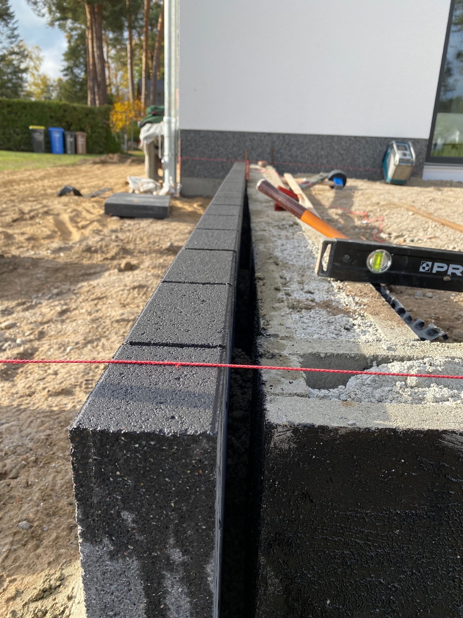 Wyrównany rząd ciemnoszarych kostek brukowych z widoczną czerwoną linką poziomującą i poziomnicą w tle, ukazujący etap budowy obrzeża chodnika.