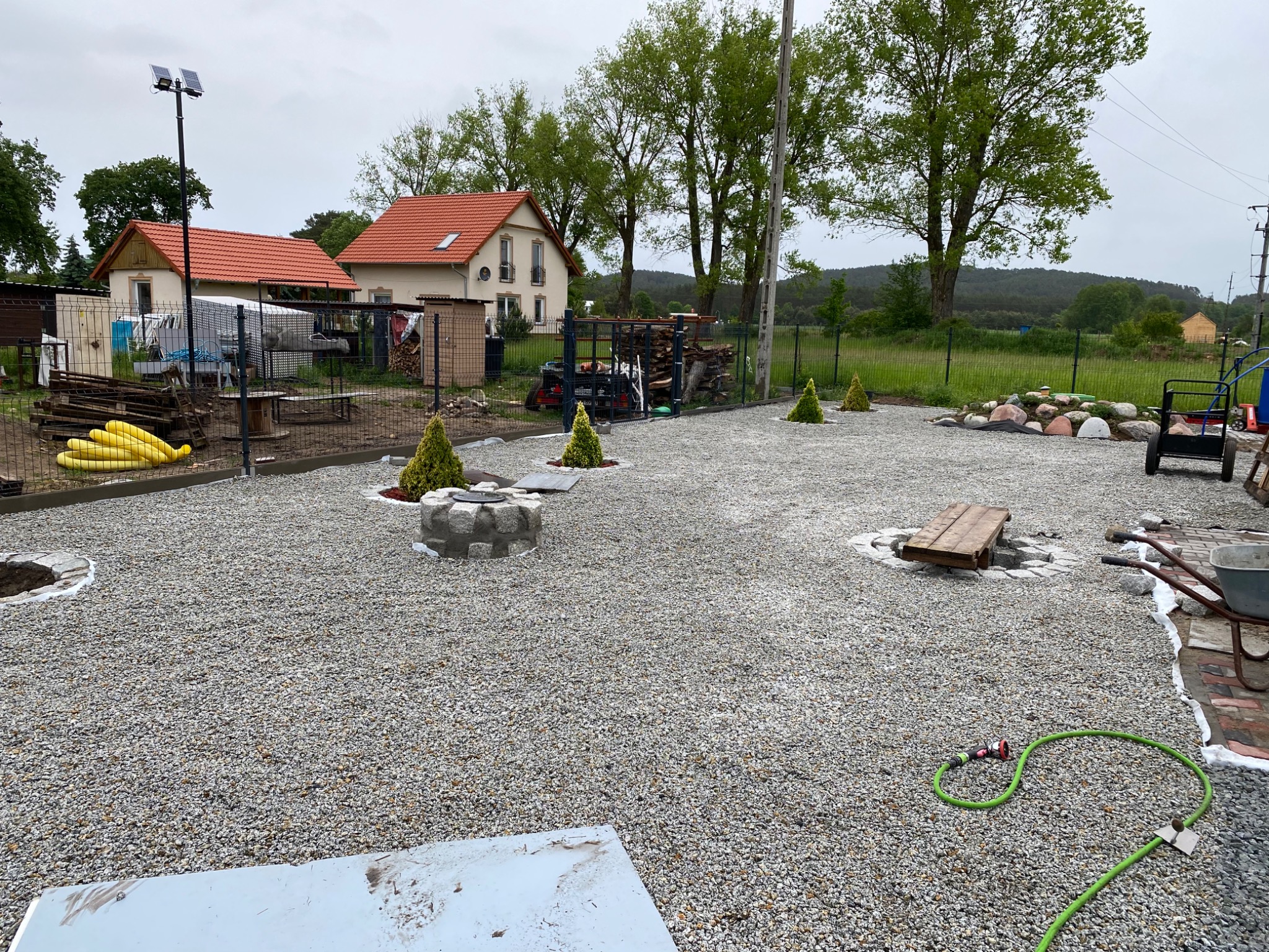 Wykończony teren wokół domu z wysypanym szarym żwirem, widoczne małe drzewka iglaste w betonowych donicach, miejsce na ognisko z kamieni oraz drewniana ławka, w tle dwa domy z pomarańczowymi dachami.
