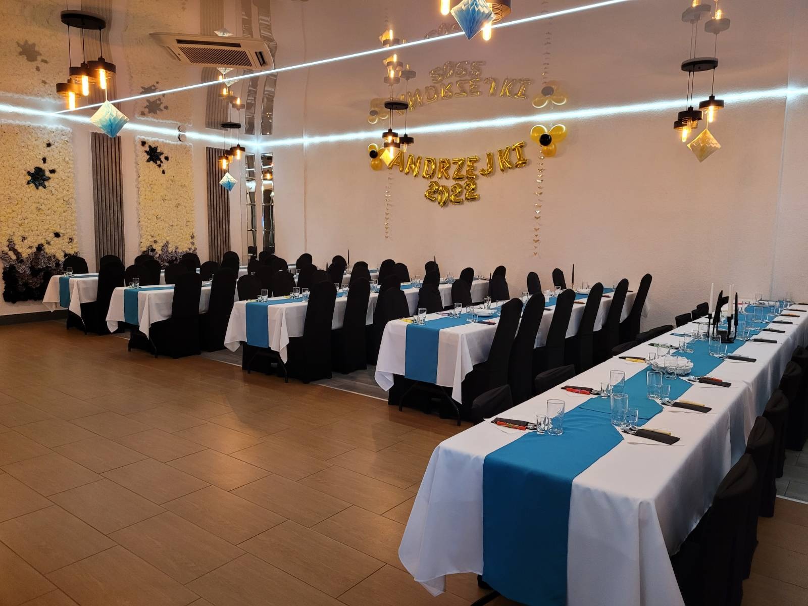 Sala bankietowa przygotowana na imprezę Andrzejkową, z długimi stołami nakrytymi białymi obrusami i niebieskimi bieżnikami, czarne pokrowce na krzesłach, dekoracje balonowe i kwiatowe na ścianach...
